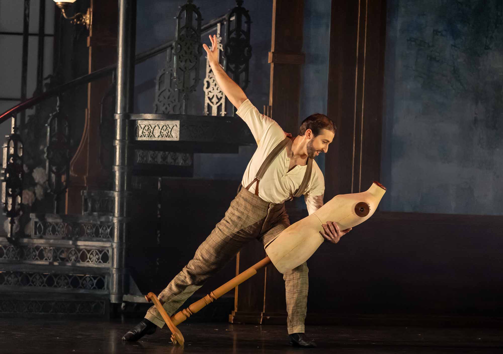 A male performer in suspenders poses dramatically on stage, holding a wooden dress form as if dancing with it. Ornate iron stairs and dim lighting create a moody theatrical atmosphere in the background.